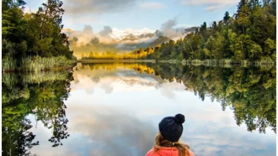 Person sitting on a dock at Lake Matheson during sunset with mountain reflections