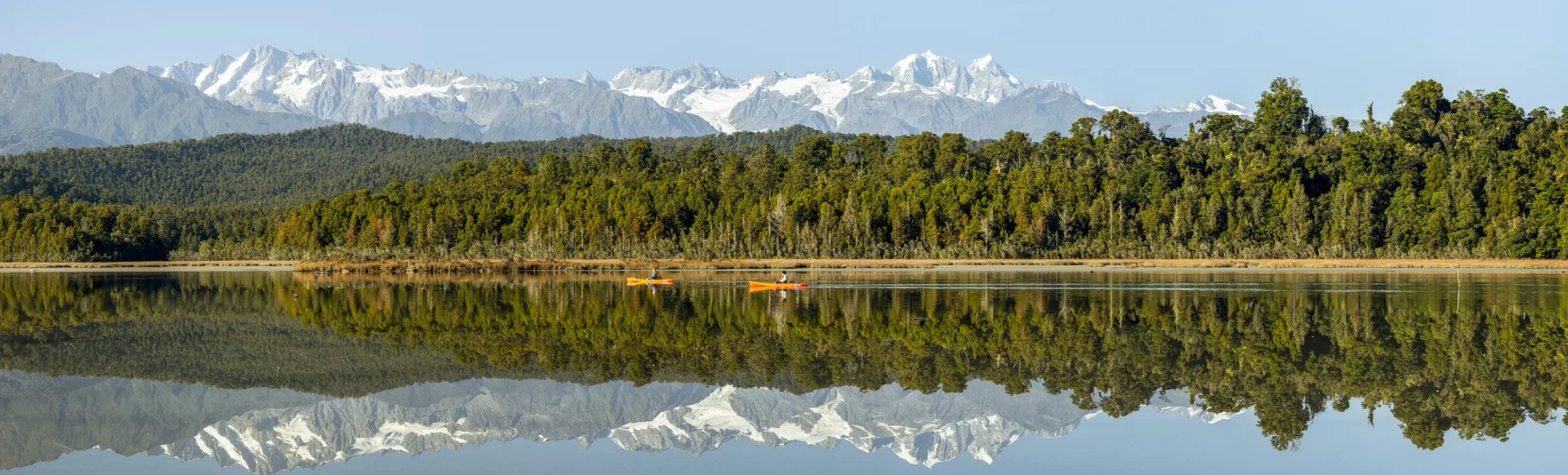 Two kayakers on Ōkārito Lagoon with snow-capped Southern Alps reflected on the water, West Coast New Zealand