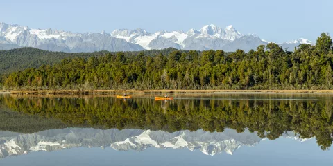 Two kayakers on Ōkārito Lagoon with snow-capped Southern Alps reflected on the water, West Coast New Zealand