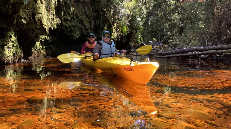 Two people kayaking on a crystal-clear creek with orange stones and lush rainforest in Ōkārito on the West Coast of New Zealand