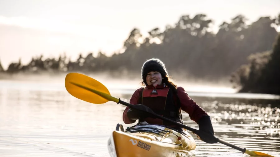 Smiling woman kayaking at sunrise on Okarito Lagoon surrounded by mist and forest