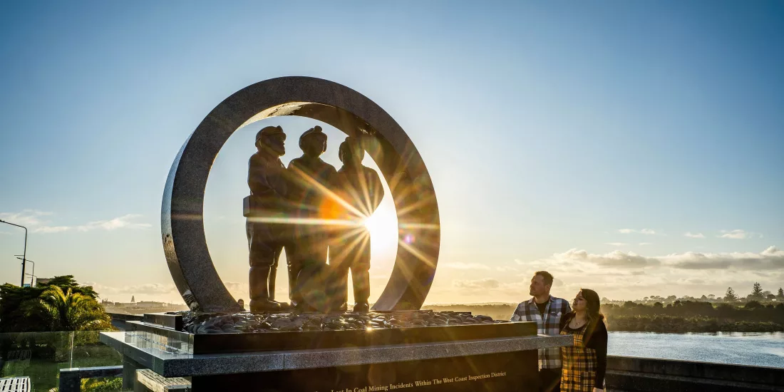 Couple standing in front of the Miners' Memorial in Greymouth at sunset, honouring those lost in West Coast coal mining incidents
