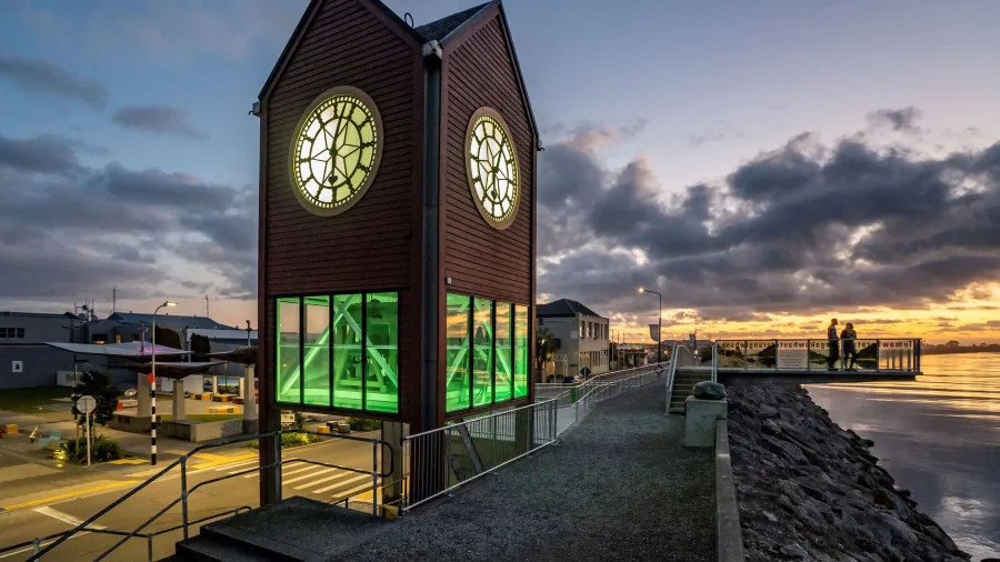 Clock tower at Māwhera Quay in Greymouth glowing green at sunset with a view of the river and boardwalk