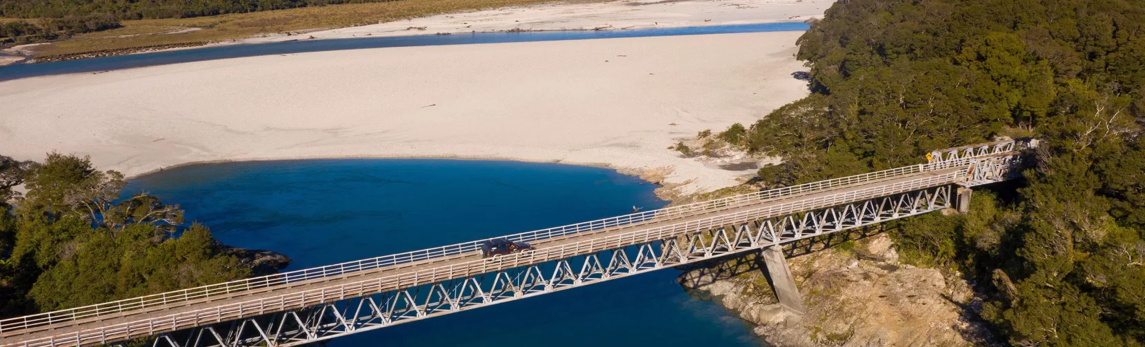 Aerial view of the bridge crossing the Arawata River with turquoise water and alpine ranges in the background