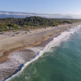 Aerial view of waves rolling onto the wide sandy beach at Ship Creek near Haast, backed by native forest and distant mountains