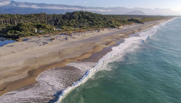 Aerial view of waves rolling onto the wide sandy beach at Ship Creek near Haast, backed by native forest and distant mountains