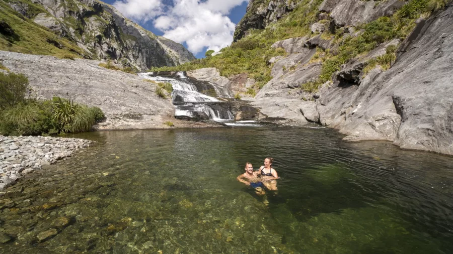 Two people swimming in a crystal-clear pool beneath a waterfall at Haast Hanging Lakes, West Coast New Zealand