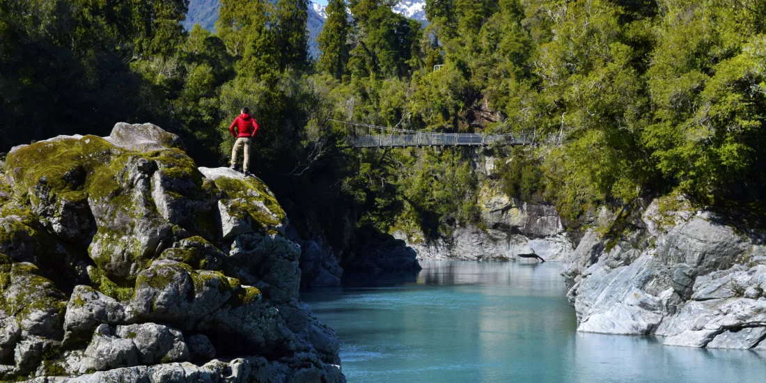 Hiker in red jacket standing on rocks beside the bright blue water of Hokitika Gorge, surrounded by native forest