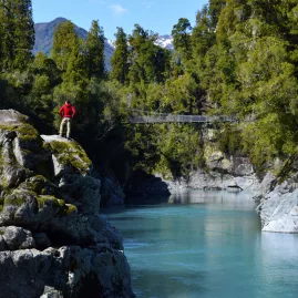 Hiker in red jacket standing on rocks beside the bright blue water of Hokitika Gorge, surrounded by native forest