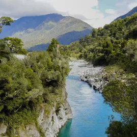 View of Hokitika Gorge with turquoise river, lush forest, and a swing bridge in the distance