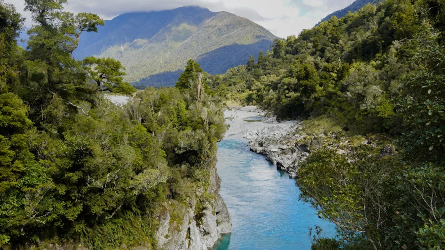 View of Hokitika Gorge with turquoise river, lush forest, and a swing bridge in the distance