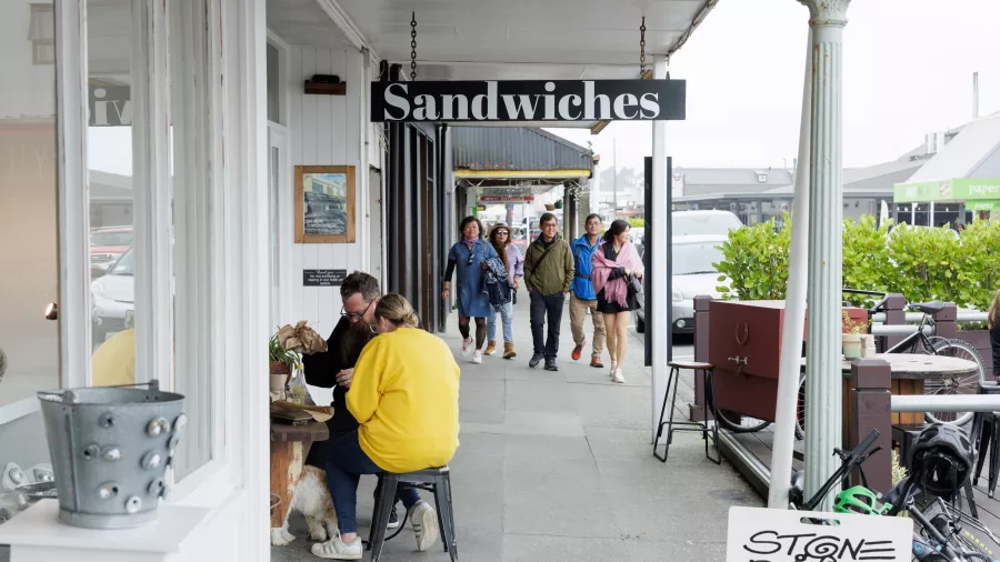 People walking and dining along a street in Hokitika town centre near a sandwich shop and stone painter studio