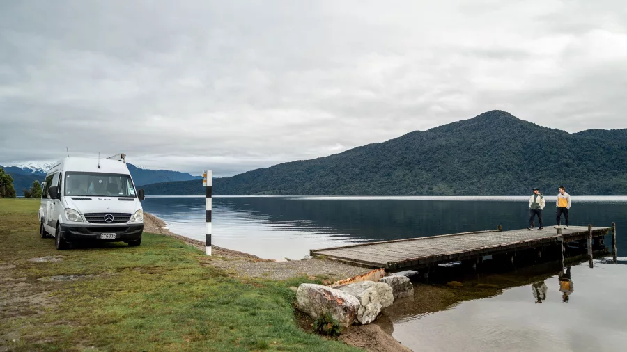Campervan parked beside the still waters of Lake Kaniere with two people walking on a jetty