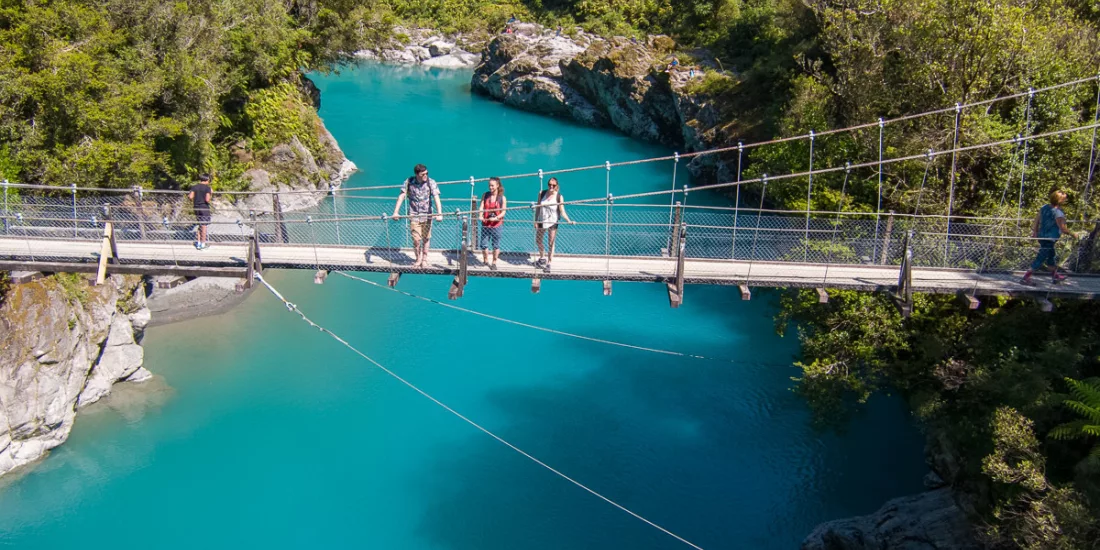 Visitors walking across the swing bridge above Hokitika Gorge’s vibrant blue river