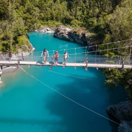 Visitors walking across the swing bridge above Hokitika Gorge’s vibrant blue river