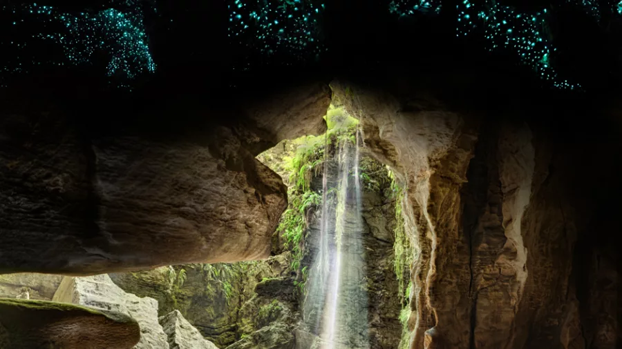 A person in a wetsuit sits in an underground cave illuminated by a natural light beam, with glowworms lighting the ceiling above during an Underworld Rafting tour in Charleston, New Zealand
