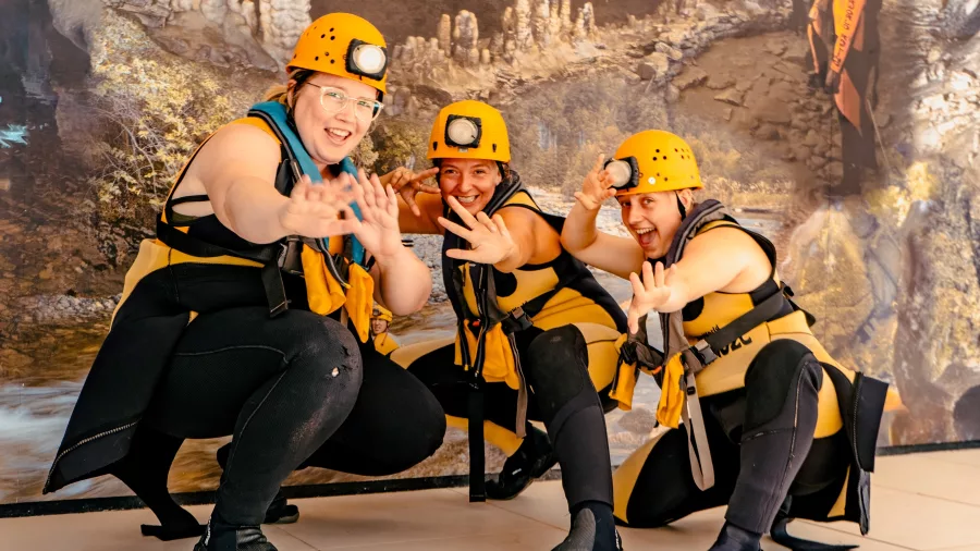 Three people in yellow wetsuits and helmets pose playfully in front of a printed cave backdrop after completing a black water rafting adventure with Underworld Adventures in Charleston, New Zealand.