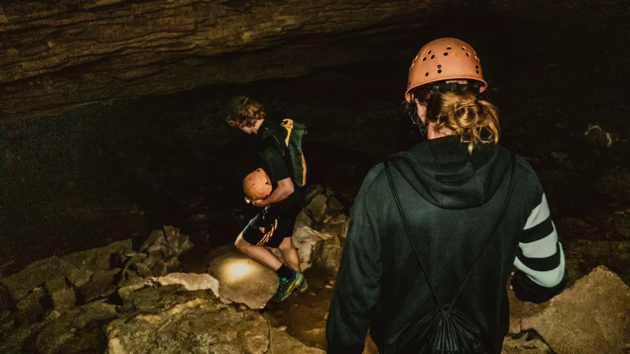 Two people wearing helmets descend into a dark cave over rocky terrain