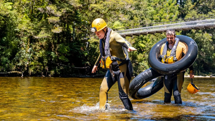 Two people in wetsuits and helmets walking through shallow river water with inner tubes after cave tubing