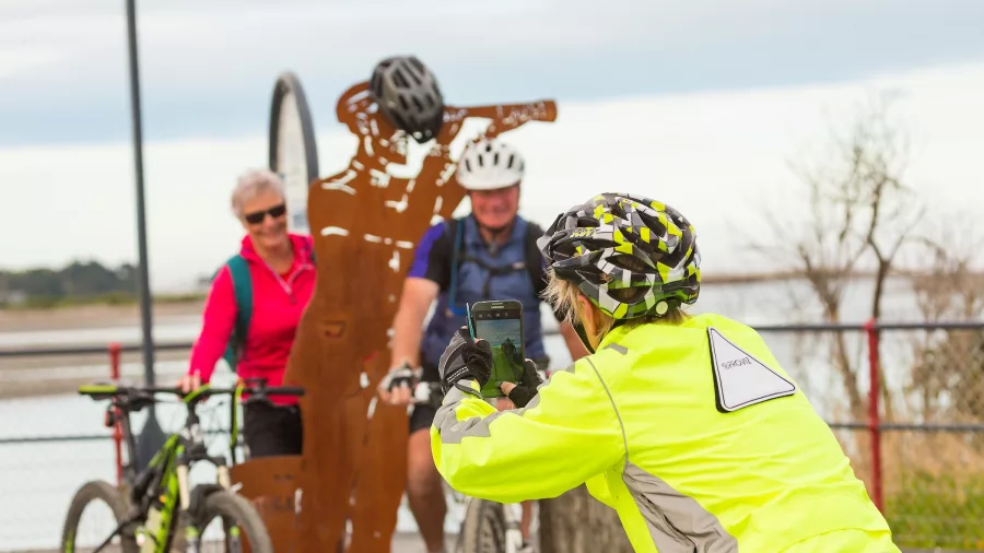 Cyclists taking a photo by a sculpture along the Hokitika River on the West Coast Wilderness Trail