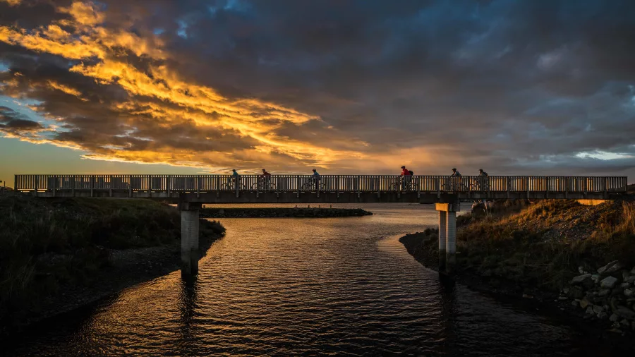 Cyclists ride across a bridge at Greymouth Lagoon during sunset on the West Coast Wilderness Trail