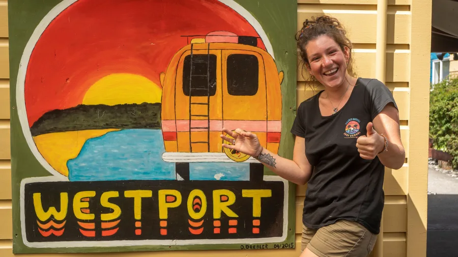 Smiling woman giving thumbs up beside a colourful Westport mural at Bazil's Hostel & Surf School